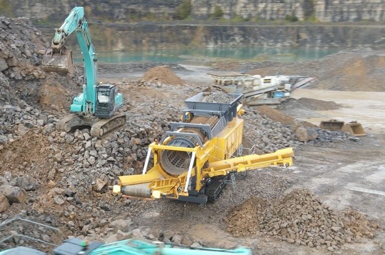ranoworks A yellow rock crusher processes stones as a green excavator loads rocks in a quarry with piles of gravel, machinery, and Ranoworks screeners visible in the background.