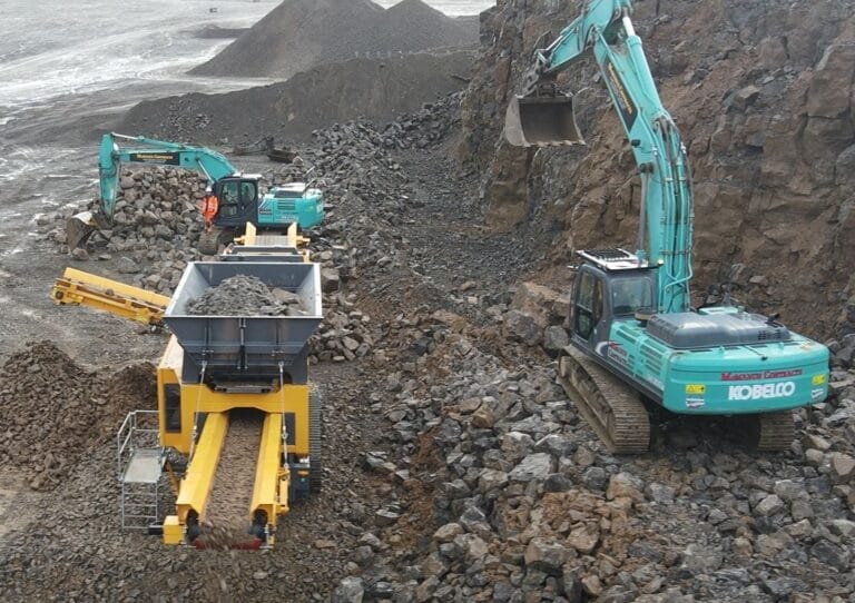ranoworks Two excavators load rocks into a mobile crusher at a rocky construction site, with crushed material exiting on conveyor belts for further processing by screening machinery.