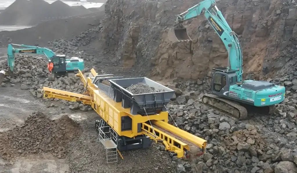 ranoworks Two excavators load rocks into a yellow mobile crusher in a quarry, with rocky cliffs behind. Ranoworks equipment like screeners and trommels efficiently process the stone piles in the background.
